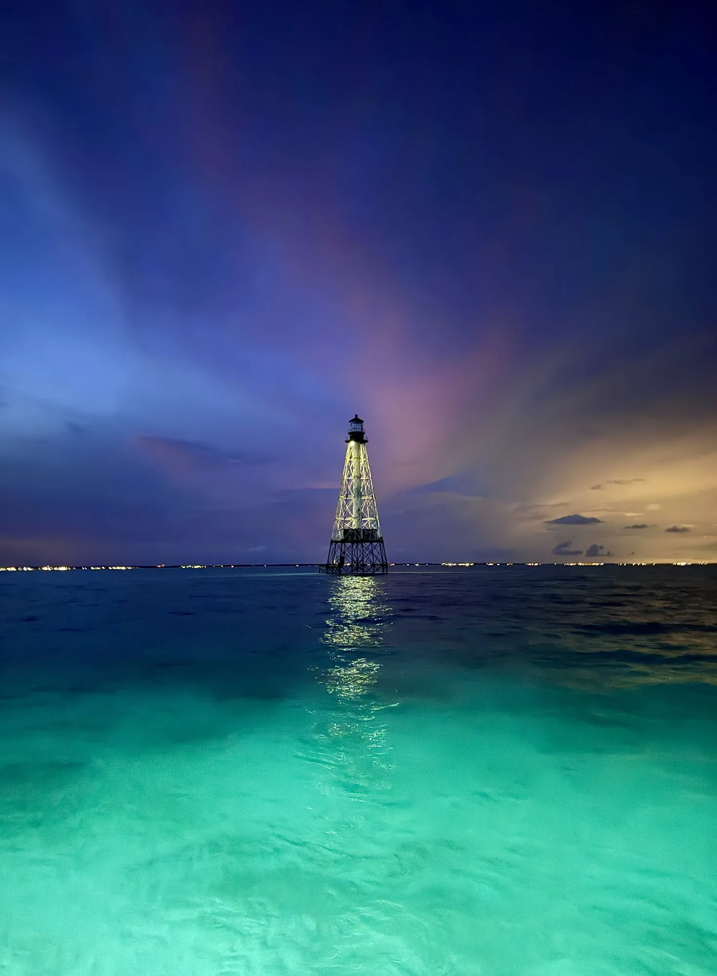 Alligator Reef Lighthouse lit up at dusk, with the boat lights turning the water turquoise