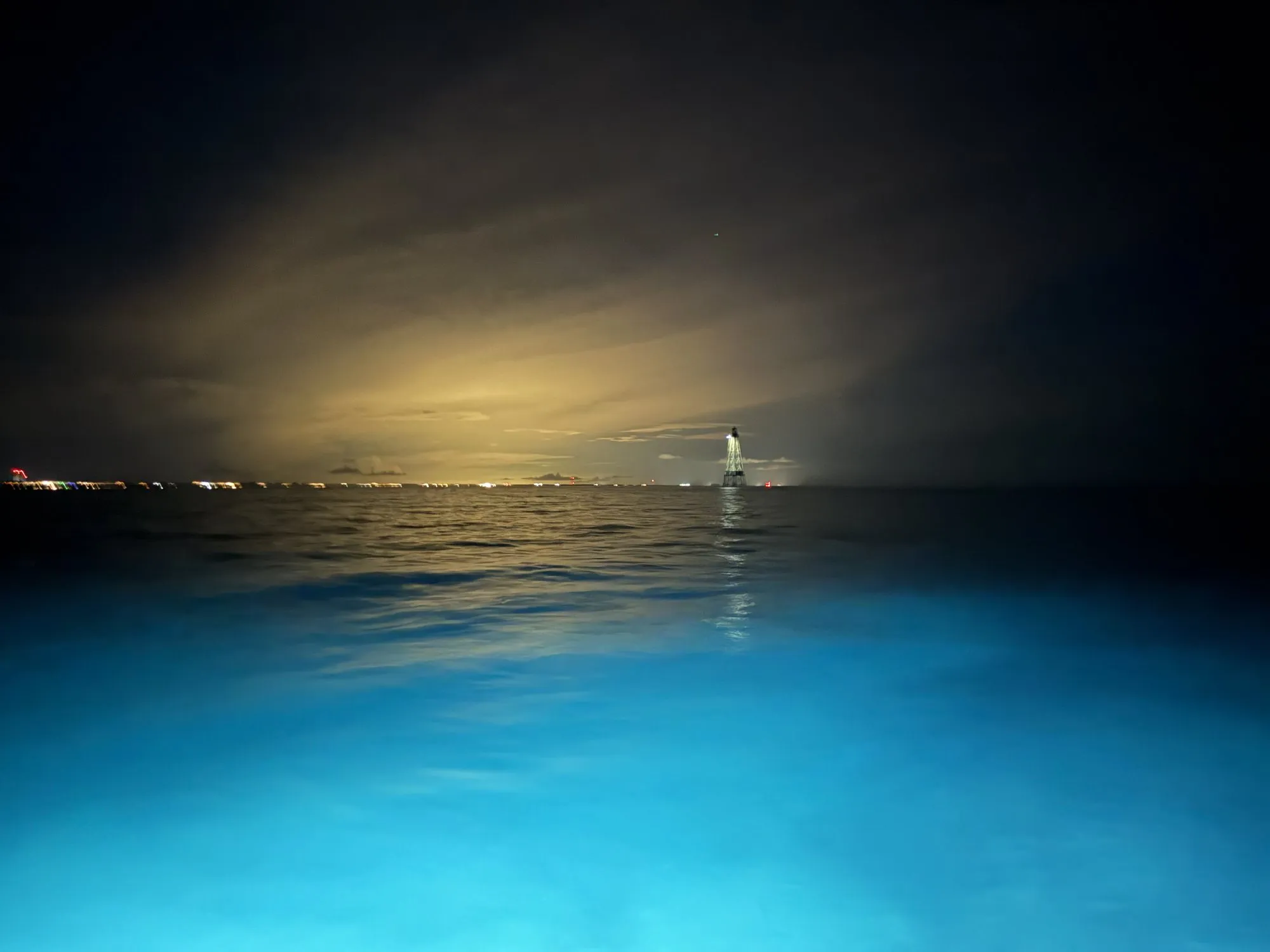 The lit lighthouse on the dark horizon after nightfall, with bright blue water around the boat