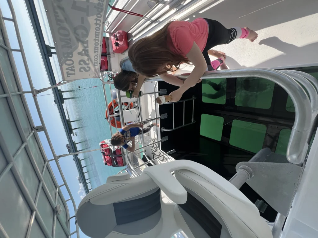 Children on a glass-bottom boat, one eating ice cream, with clear ocean and pier.